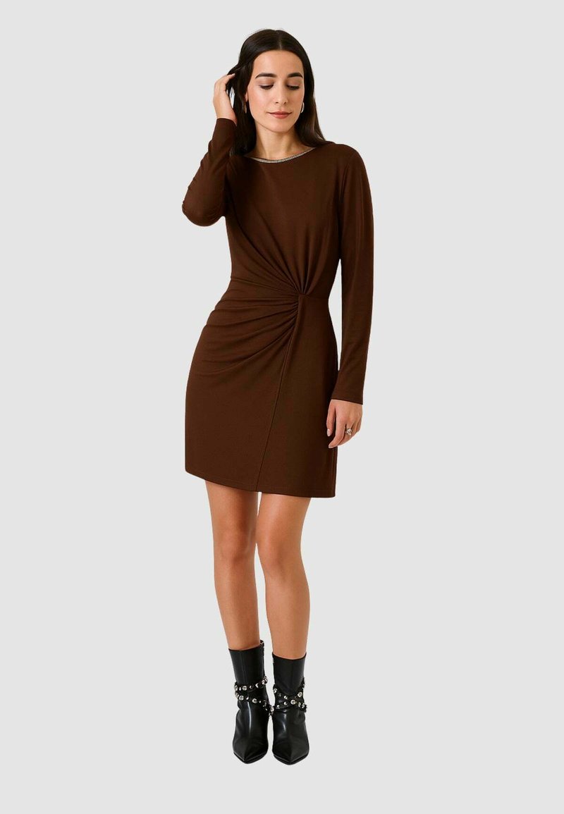 Vestito marrone a maniche lunghe con dettaglio nodo in vita, tessuto liscio e silhouette dritta. Abbinato a stivaletti neri.