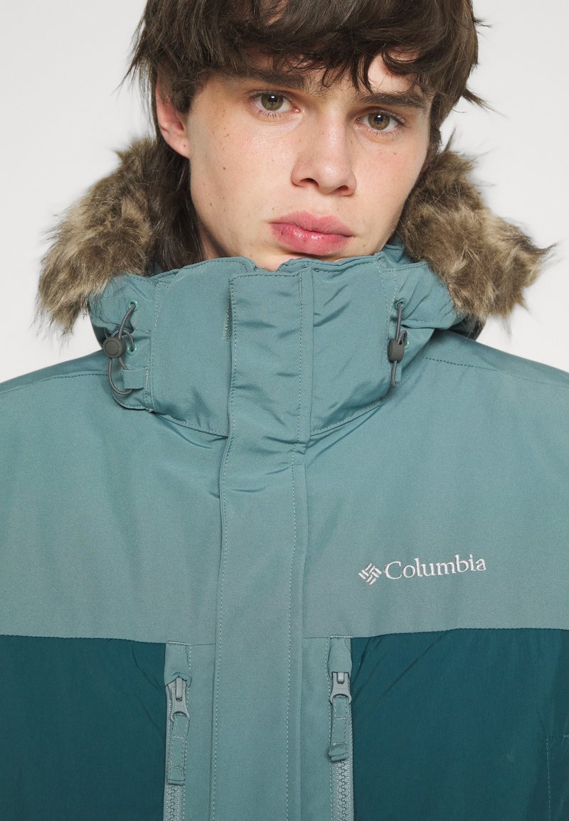 Veste Columbia teal avec une capuche doublée de fausse fourrure, zips sur le devant et logo sur la poitrine. Matériau lisse, design rembourré.