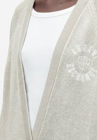 Gros plan d'une personne portant un haut blanc côtelé et un cardigan beige avec le texte brodé « Albion Exchange » et un emblème en forme de bouclier.