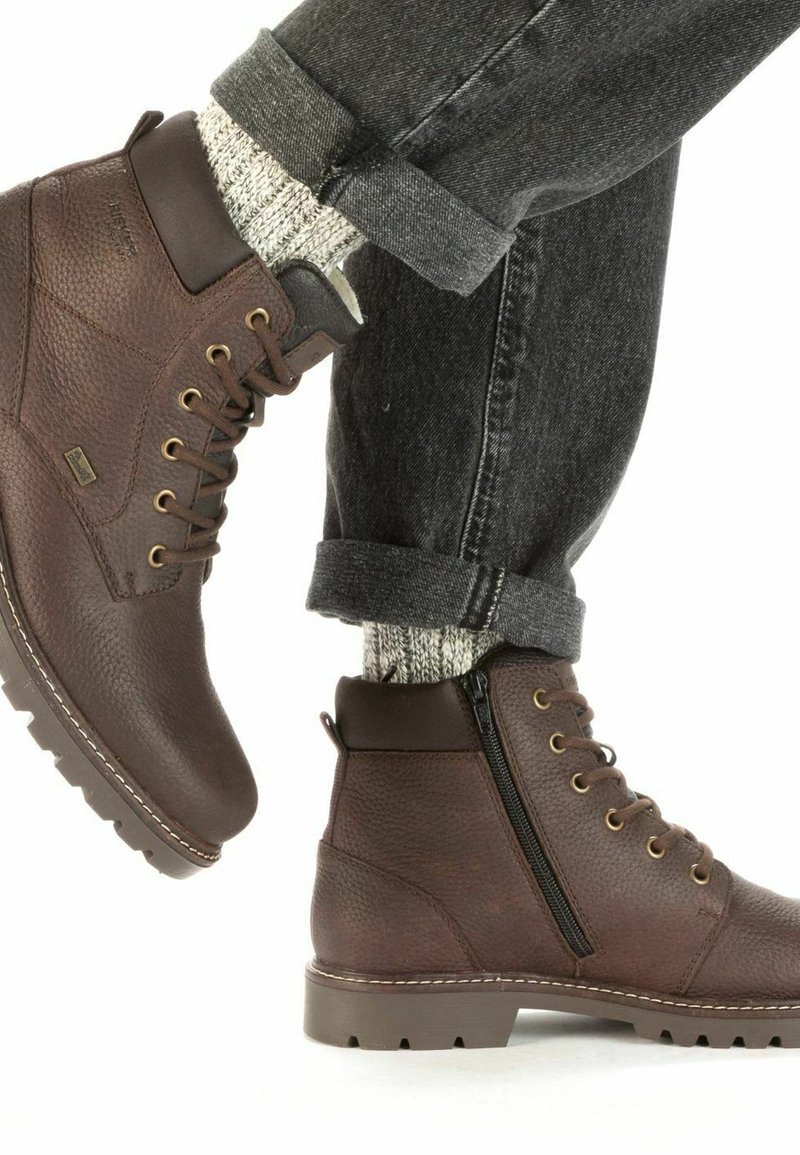 Botas de cuero marrón hasta el tobillo con acabado texturizado, cordones en el frente, cremallera lateral y suela de goma. Combinadas con unos vaqueros oscuros doblados y calcetines de punto.