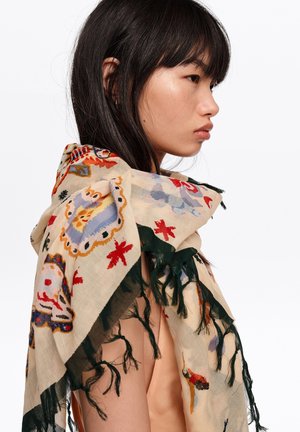 Młoda kobieta z długimi ciemnymi włosami, ubrana w beżowy szalik z kolorowymi wzorami i czarnymi frędzlami, pokazana z profilu na białym tle.