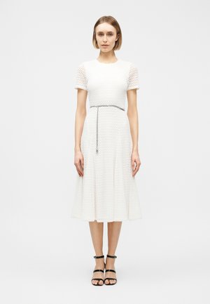 Kvinne som står iført hvit teksturert midikjole med korte ermer og et tynt kjede belte, kombinert med svarte sandaler med ankelstropp.