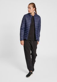 Giacca puffer blu navale con colletto alto, design trapuntato, chiusura con zip, indossata sopra un maglione nero e pantaloni della tuta neri, completata da sneakers nere.