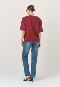 Bordová oversized tričko s krátkými rukávy, spárované s modrými rovnými džíny a černými podpatky, viděná ze zadní strany. Hladká textura.