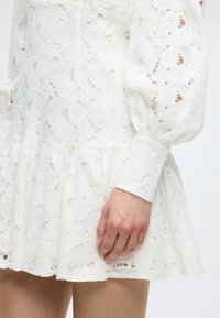 Λευκό δαντελένιο φόρεμα με μακριά φουσκωτά μανίκια και βολάν στο τελείωμα, φορεμένο από ένα άτομο με το χέρι να ακουμπά κοντά στη φούστα.