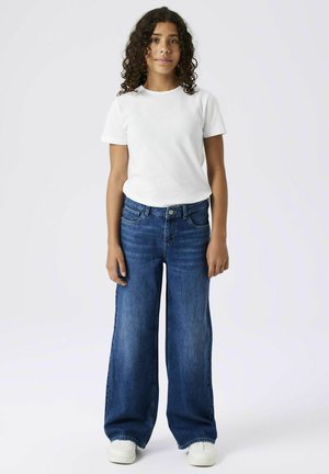 White t-shirt with short sleeves, paired with blue wide-leg jeans and white sneakers. Smooth fabric and relaxed fit, featuring pockets.