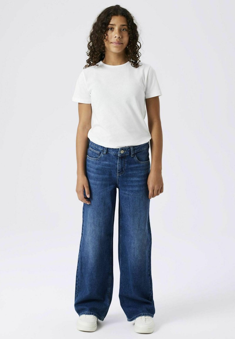 T-shirt blanc à manches courtes, associé à un jean large bleu et des baskets blanches. Tissu lisse et coupe décontractée, avec poches.