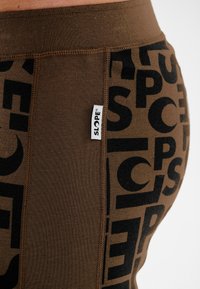 Braune Leggings mit einem auffälligen schwarzen Grafikmuster und einem kleinen weißen Etikett mit "SLOPE" am Bund. Mischung aus glatten Texturen.