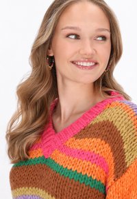 Maglione lavorato a maglia colorato con scollo a V e strisce nei colori rosa, arancione, marrone, verde e lavanda. La modella indossa orecchini a cerchio con perline.