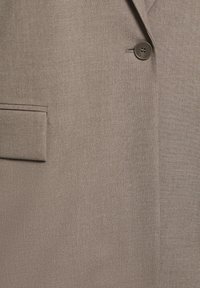 Veste de costume en tissu brun clair avec un bouton foncé unique et une poche à rabat sur le côté gauche.