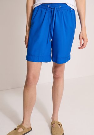 Person iført klare blå knælange shorts med snøre, hvid skjorte og lysebrune hjemmesko, der står indendørs mod en ensfarvet baggrund.