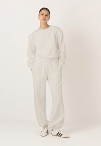 Világosszürke pulóver és hozzá illő melegítőnadrág, mindkettő puha anyagból készült, lazán szabott, bordás mandzsettákkal és oldalsó zsebekkel. Csíkos sportcipő.