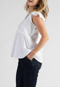 Weiße Baumwollbluse mit Lochstickerei, kurzen Rüschenschultern und lockerer Passform, kombiniert mit dunklen Denim-Jeans.