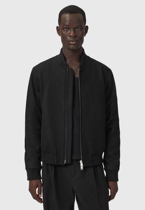 Mann trägt eine schwarze Zip-up-Jacke, ein schwarzes Hemd und schwarze Hose und steht vor einem einfarbigen hellgrauen Hintergrund.