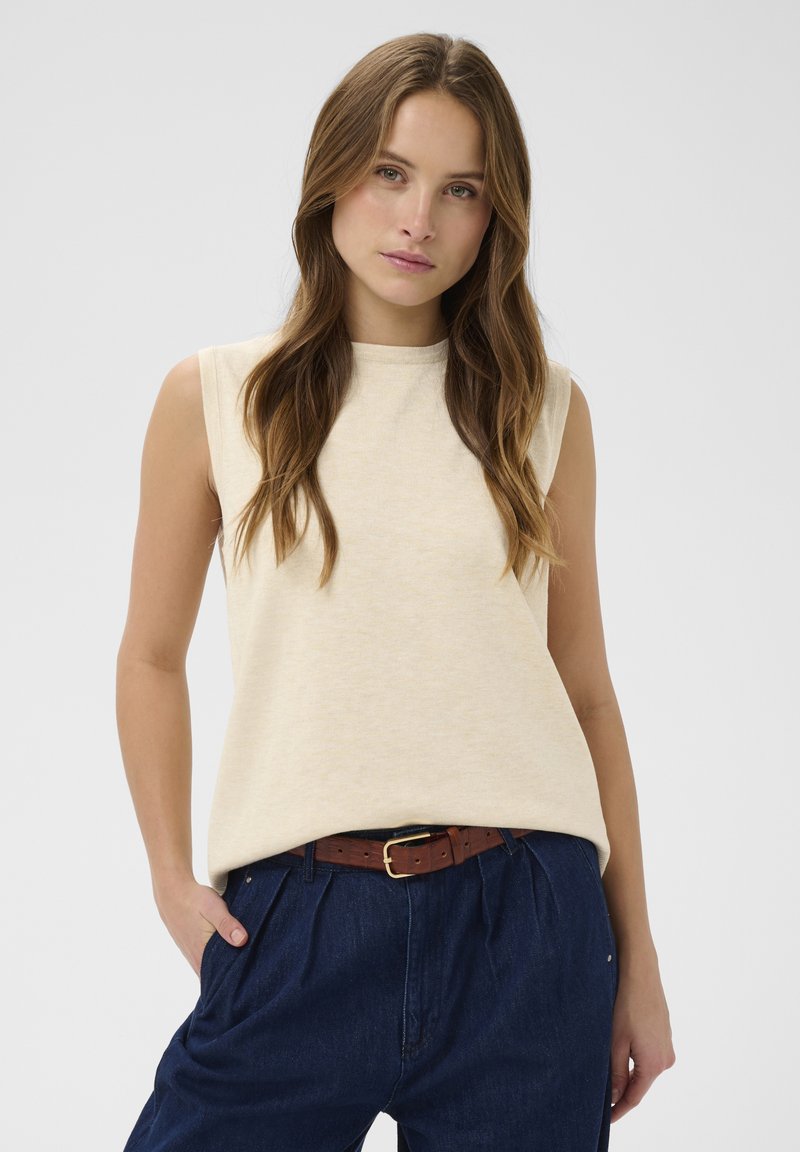 Bežinė be rankovių palaidinė, pagaminta iš minkštos, tekstūruotos medžiagos. Apykaklė su apvaliu iškirpimu, laisvo kirpimo, derinama su tamsiai mėlynomis raukšlėtomis kelnių ir rudą diržu.