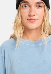 Ανοιχτό μπλε μακρυμάνικη μπλούζα με στρογγυλή λαιμόκοψη, απαλό ύφασμα και κεντημένο λογότυπο στο στήθος. Φορεμένη με μαύρο σκουφί.