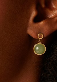Gouden oorbellen met een ronde groene steen die hangt aan een ronde hoepel. De steen heeft een gladde textuur en een glanzende afwerking.