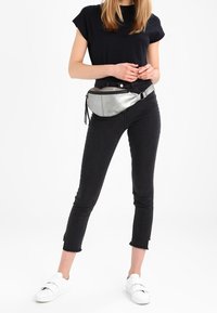 Hopeinen metallinen vyölaukku, joka on sijoitettu vyötärölle mustien kireiden farkkujen päälle, yhdistetty valkoisiin lenkkkenkiin ja lyhythiharaiseen mustaan paitaan.