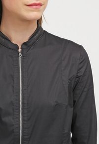 Svart zip-up jacka med ståkrage, med en mjuk, glänsande textur, kontrasterande läderdetaljer och synliga sömmar.