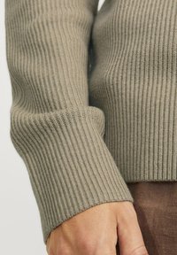 Pulover bej cu dungi, având mâneci lungi cu manșete groase, realizat dintr-un material tricotat moale cu textură. Prim-planul arată o mână sprijinită pe o țesătură maro.
