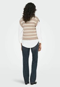 Henkilö, jolla on kiharat hiukset, pukeutuneena beige-valkoiseen raitapaitaan ilman hihoja valkoisen paidan päällä ja tummansinisiin farkkuihin, seisoo tasaisella taustalla.