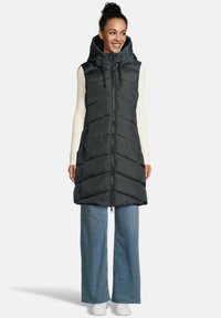 Vrouw in lange zwarte gewatteerde mouwloze jas met capuchon over een witte geribbelde lange mouw top, blauwe wijde jeans en witte sneakers, lachend.