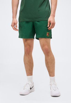 Person trägt grüne Sportshorts mit Logo, weiße Socken und weiße Nike-Sneakers, steht vor einem einfarbigen Hintergrund.