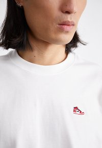 T-shirt in cotone bianco con un logo di scarpa ricamato rosso e nero sul lato sinistro del petto. Collo rotondo e maniche corte.