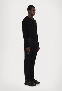 Black long-sleeve polo shirt with a relaxed fit, paired with black tailored trousers and black sneakers. Subtle branding on the shirt.