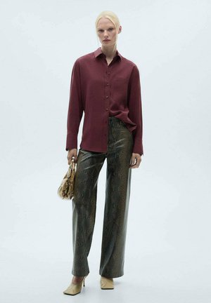 Bordeaux kleurige blouse met knopen gecombineerd met hoge taille, groene slangenpatroon wijde broek. Model houdt een gemusterde handtas vast en draagt beige hakken.
