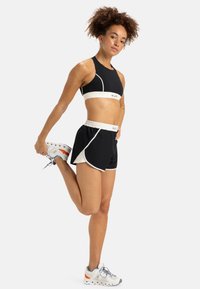 Zwarte sportbeha met witte accenten en logo, gecombineerd met zwarte shorts met witte bies en een koord in de taille, gedragen met grijze sneakers.