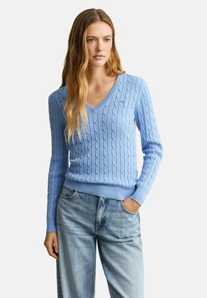 Frau mit langem, welligem Haar, die einen hellblauen, kabelgestrickten V-Ausschnittpullover und lockere hellblaue Jeans trägt, mit einer Hand in der Tasche stehend.