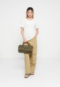 Woman stands holding olive green handbag, wearing white t-shirt, beige cargo pants, and patterned sneakers against plain white background.