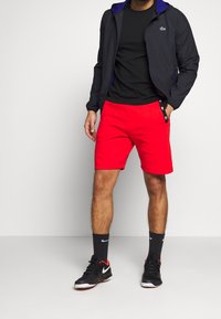 Röda atletiska shorts med sidofickor, svart lättviktsjacka med blå huva, och svarta sneakers med röda detaljer; svart t-shirt under.