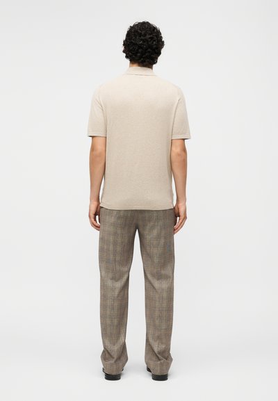 Άνδρας με σγουρά μαλλιά στέκεται με την πλάτη, φοράει ένα μπεζ κοντομάνικο πόλο και γκρι καρό παντελόνι με μαύρα παπούτσια σε απλό φόντο.