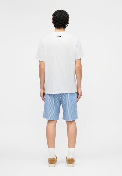 Άνδρας με σγουρά μαλλιά που φοράει λευκό μπλουζάκι MSGM, μπλε και λευκές ριγέ βερμούδες, λευκές κάλτσες και καφέ αθλητικά παπούτσια, στέκεται γυρισμένος ανάποδα.