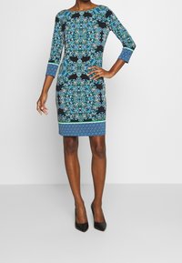 Kortärmad klänning i blått och svart med intrikata blommönster och geometriska mönster, med en figurnära siluett och rund halsringning.