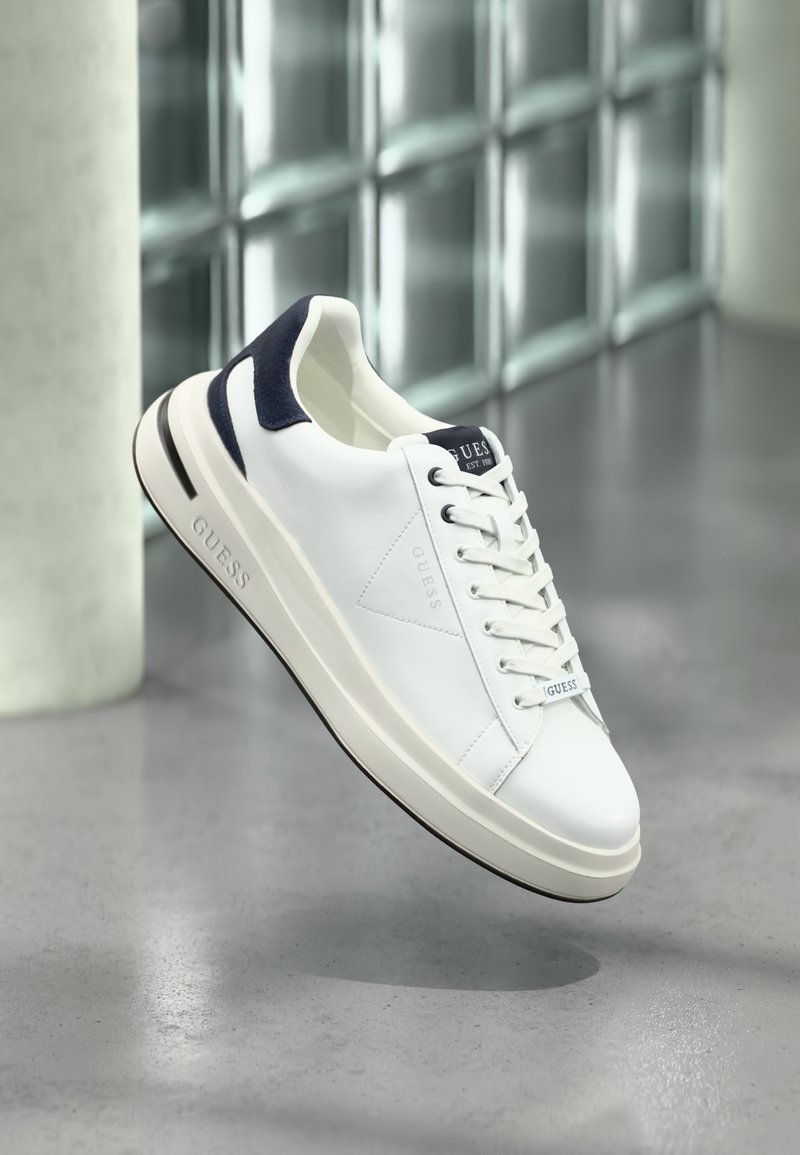 Sapatilhas brancas em couro com detalhes em azul marinho, ponta arredondada, calcanhar texturizado e sola plana em borracha, apresentando a marca "GUESS" na lateral.