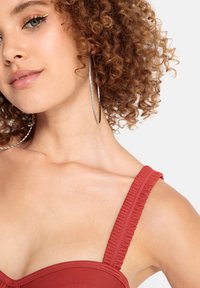 Bordo maudymosi palaidinė su raukiniais ant pečių ir prigludusia forma. Matomi garbanoti plaukai ir dideli sidabriniai auskarai žiede. Lygi odos spalva.