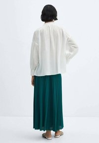 Witte, losvallende blouse met een geraffineerde rug; gecombineerd met een lange, teal kleurige plissé rok. Gecombineerd met platte sandalen. Eenvoudige, lichte stof.