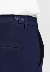 Gros plan de la taille d'un pantalon bleu marine avec une fermeture à double bouton et une petite poche arrière fendue, associé à un haut blanc à côtes.