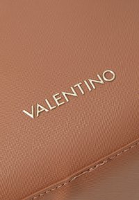 Hnědý texturovaný materiál s jemným vzorem; výrazné zlaté logo "VALENTINO" s oblými písmeny. Čistý, minimalistický design.