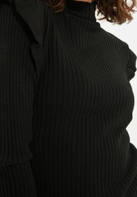 Czarny sweter z prążkowanego materiału z wysokim kołnierzem i falbanami na lewym ramieniu. Strukturalna tkanina dodaje wizualnego zainteresowania.