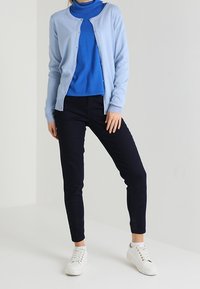 Jasnoniebieski kardigan noszony na niebieskim golfie, w połączeniu z granatowymi spodniami i białymi sneakerami. Kardigan ma guziki i miękką fakturę.