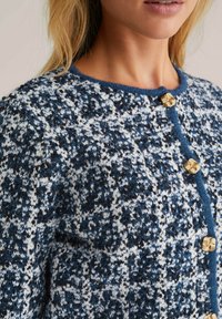 Knitted cardigan featuring a blue and white speckled pattern, gold-tone decorative buttons, and a crew neckline with a contrasting trim.