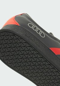 Baskets noires et rouges avec une tige lisse, une semelle en caoutchouc texturée, et un logo Audi bien visible. Présentent un accent rouge contrastant sur le côté.