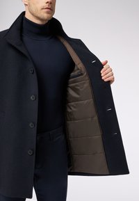 Cappotto di lana blu navy con colletto a punta, design a un solo petto e chiusura con bottone. All'interno presenta un rivestimento imbottito marrone con tasca.