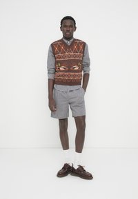 Καφετί γιλέκο με σχέδιο πάνω από πουκάμισο με καρό μοτίβο, συνδυασμένο με σορτς με καρό σχέδιο και καφέ παπούτσια. Οι λευκές κάλτσες ολοκληρώνουν το σύνολο.