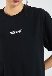 Černé bavlněné tričko s malým bílým vyšitým textem v japonštině, kulatým výstřihem a krátkými rukávy. Hladká textura, volný střih.