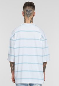 Tricou alb cu dungi orizontale de culoare albastru deschis, mâneci scurte și o croială lejeră. Prezintă un guler rotund și tatuaje vizibile pe brațe.